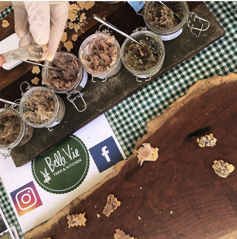 A hand with a gloved finger holds a spoon above jars of various meat spreads displayed on a wooden board, alongside a logo for Belle Vie Farm & Kitchen, set on a green checkered tablecloth.