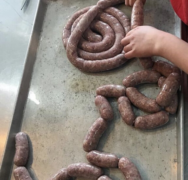 Person shaping freshly made sausages on a metal tray, showcasing various lengths and twists of the sausage links. The image highlights the art of sausage making, with a focus on the texture and color of the raw sausage meat.