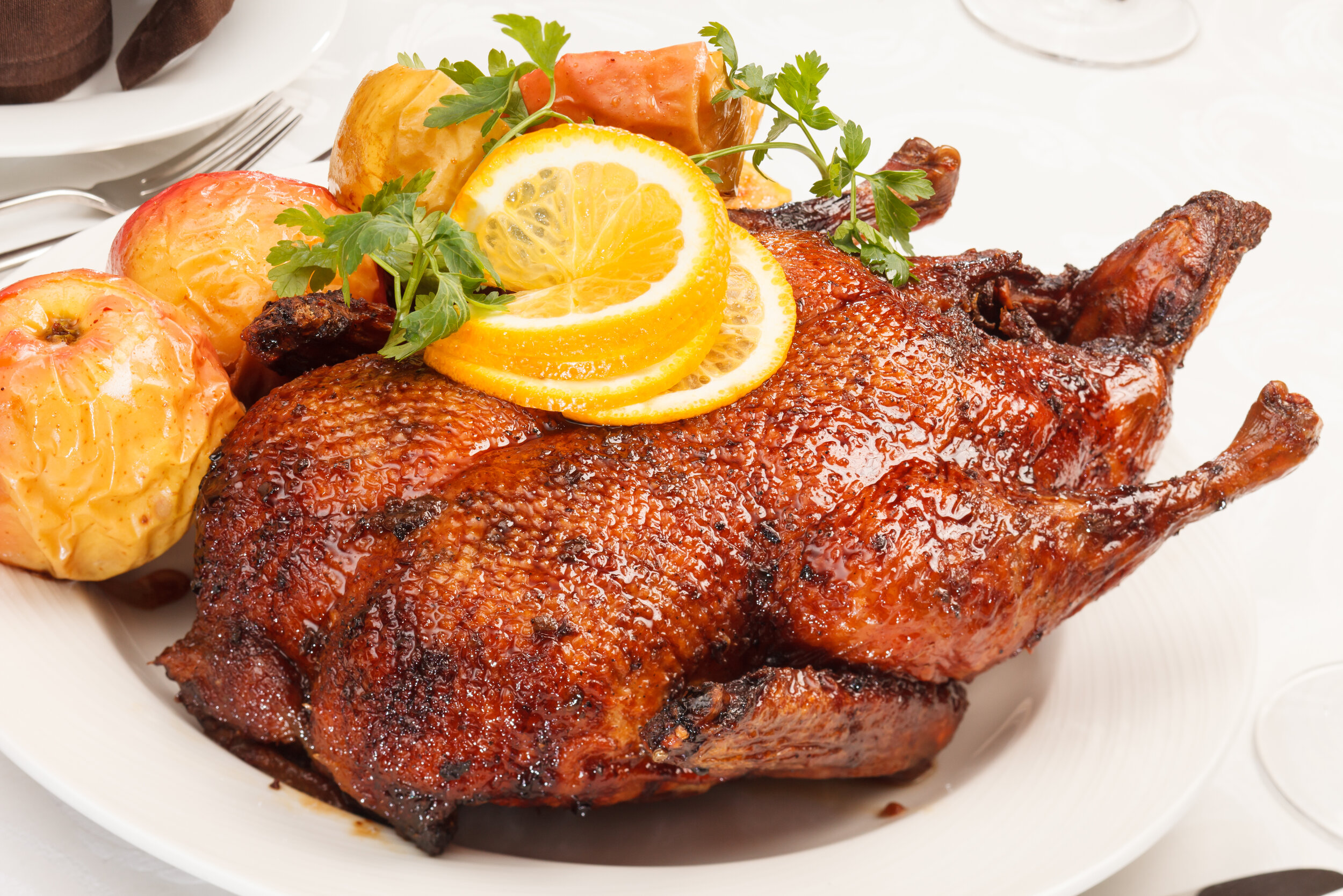Roasted duck garnished with orange slices and fresh herbs, served with baked apples on a white plate, showcasing a delicious and festive dish ideal for special occasions.