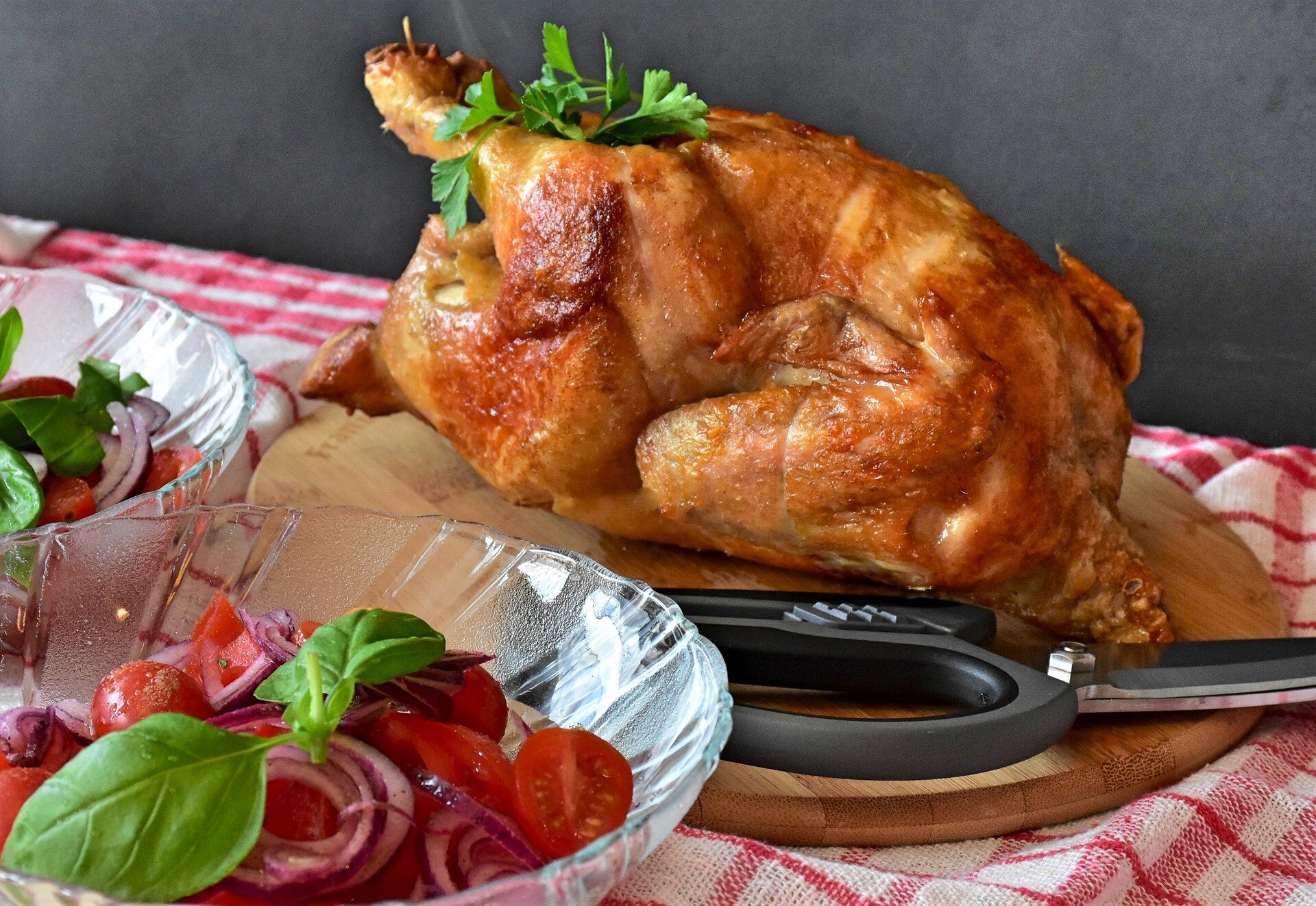 Roasted whole chicken garnished with fresh parsley on a wooden cutting board, accompanied by two bowls of fresh tomato and onion salad with basil leaves, set against a checkered tablecloth.
